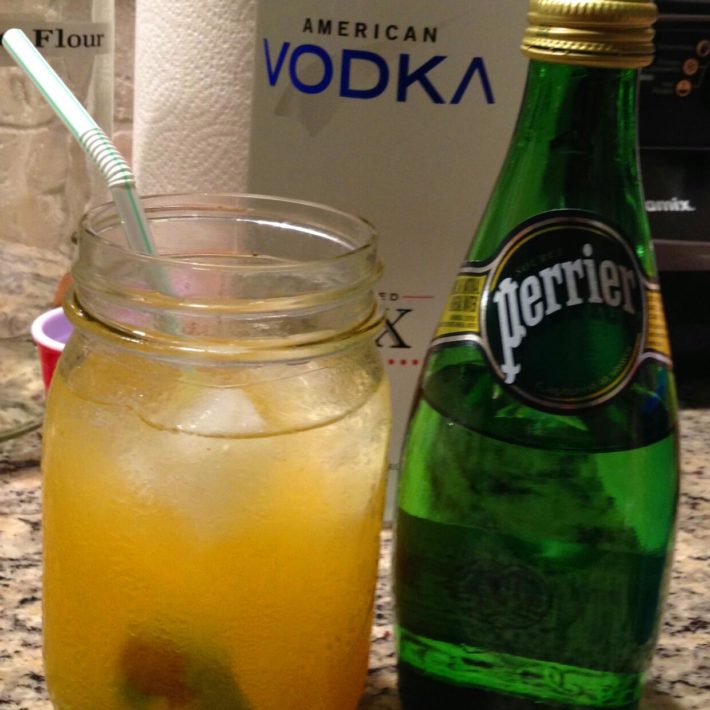 Tall Nani Recipe with in a glass with a lime and a straw along side a bottle of perrier